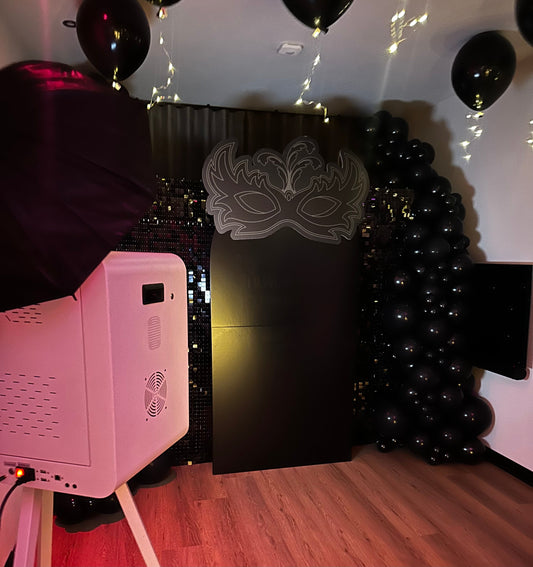 Fotobooth-opstelling met een zwarte ballonboog, glinsterende achtergrond, grote maskerdecoratie en sfeerverlichting, klaar voor gebruik tijdens een gemaskerd bal of themafeest.